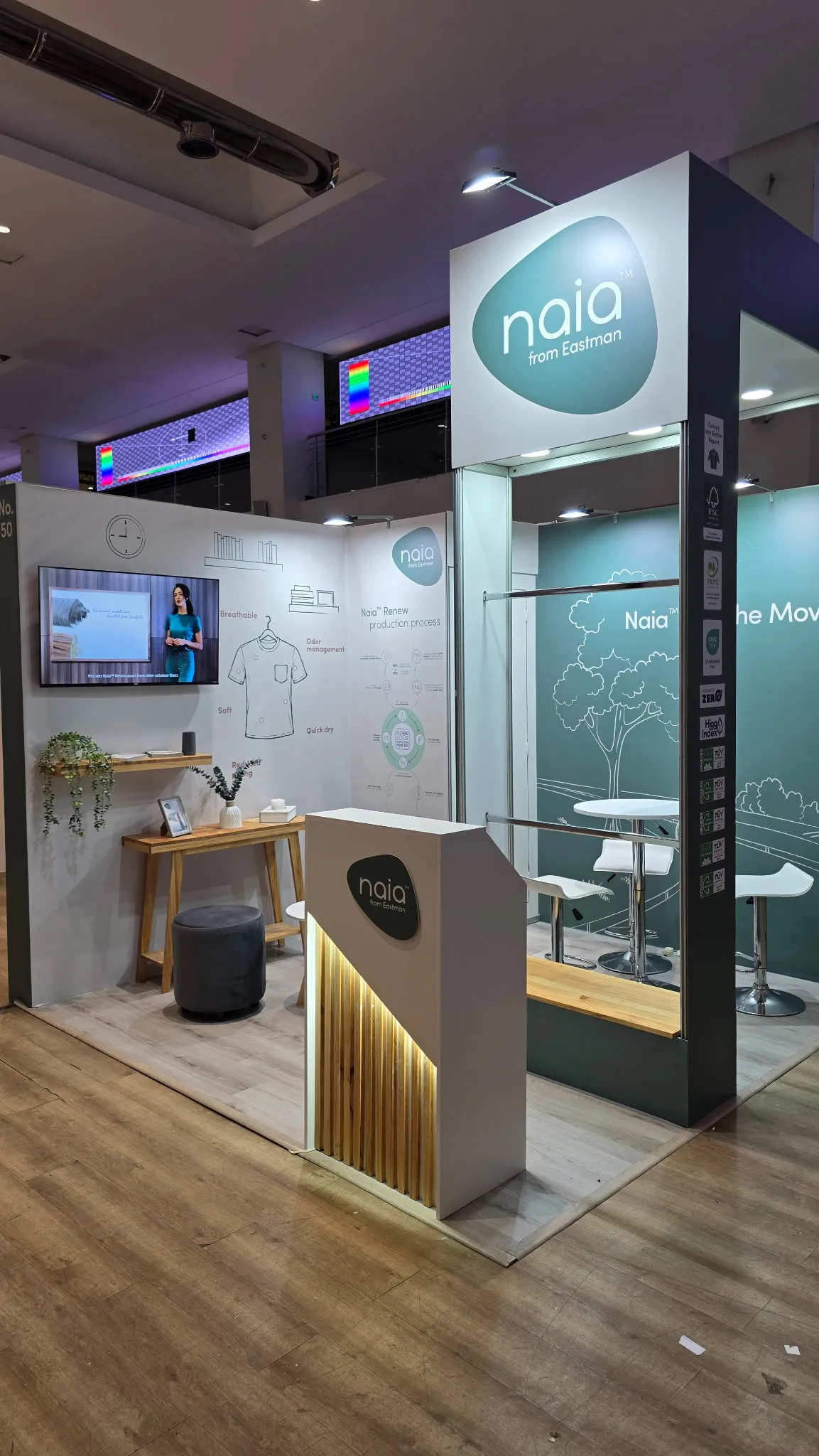 Naia booth viewed from the entrance, with reception counter, display tower, and illustrated walls.